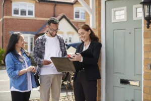 A female estate agent in a black blazer smiles while showing documents in a leather folder to a young couple standing outside a modern brick house with a light green front door.