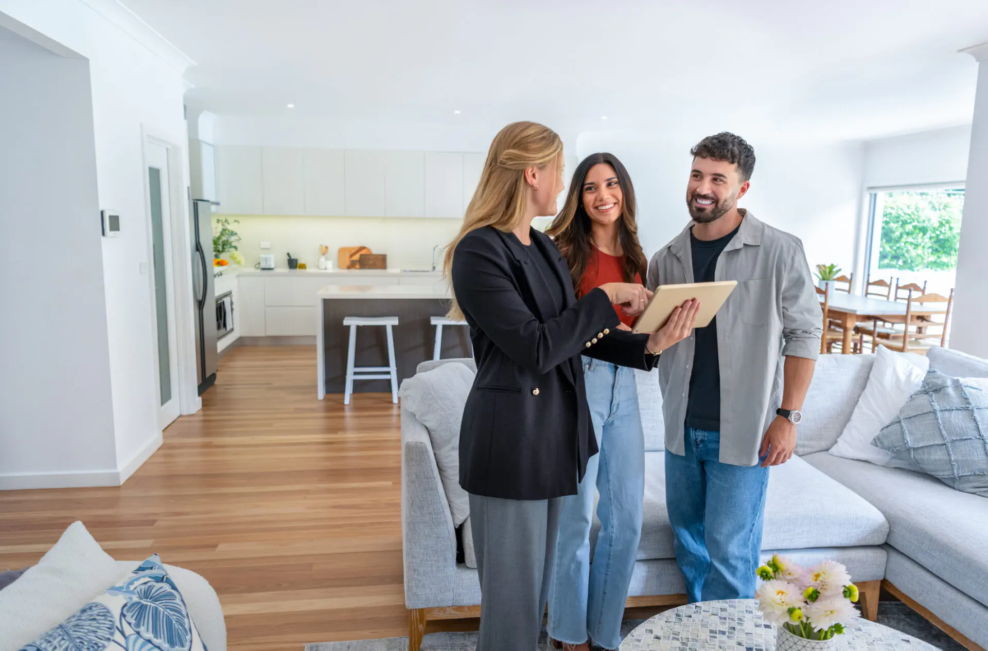 A real estate professional in a black blazer shows information on a tablet to a smiling young couple in the living room of a modern, open-plan home.